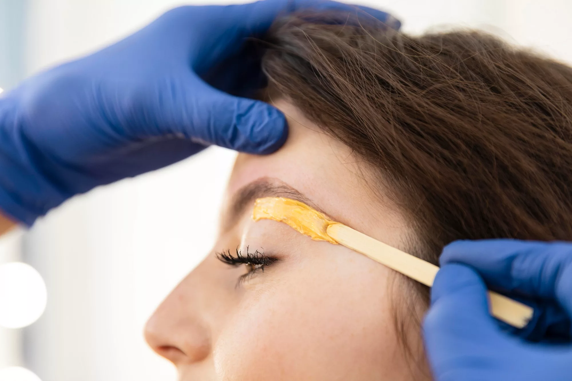 Manos con guantes poniendo cera en las cejas para hacer depilación